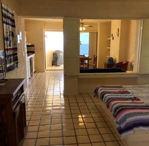 A room with a tiled floor, a bed with a patterned blanket, a kitchenette, a dining area with a table and chairs, and a view of the sea through a window. - Cozy Studio condo in Terrasol with beautiful ocean view. (Cabo san Lucas)