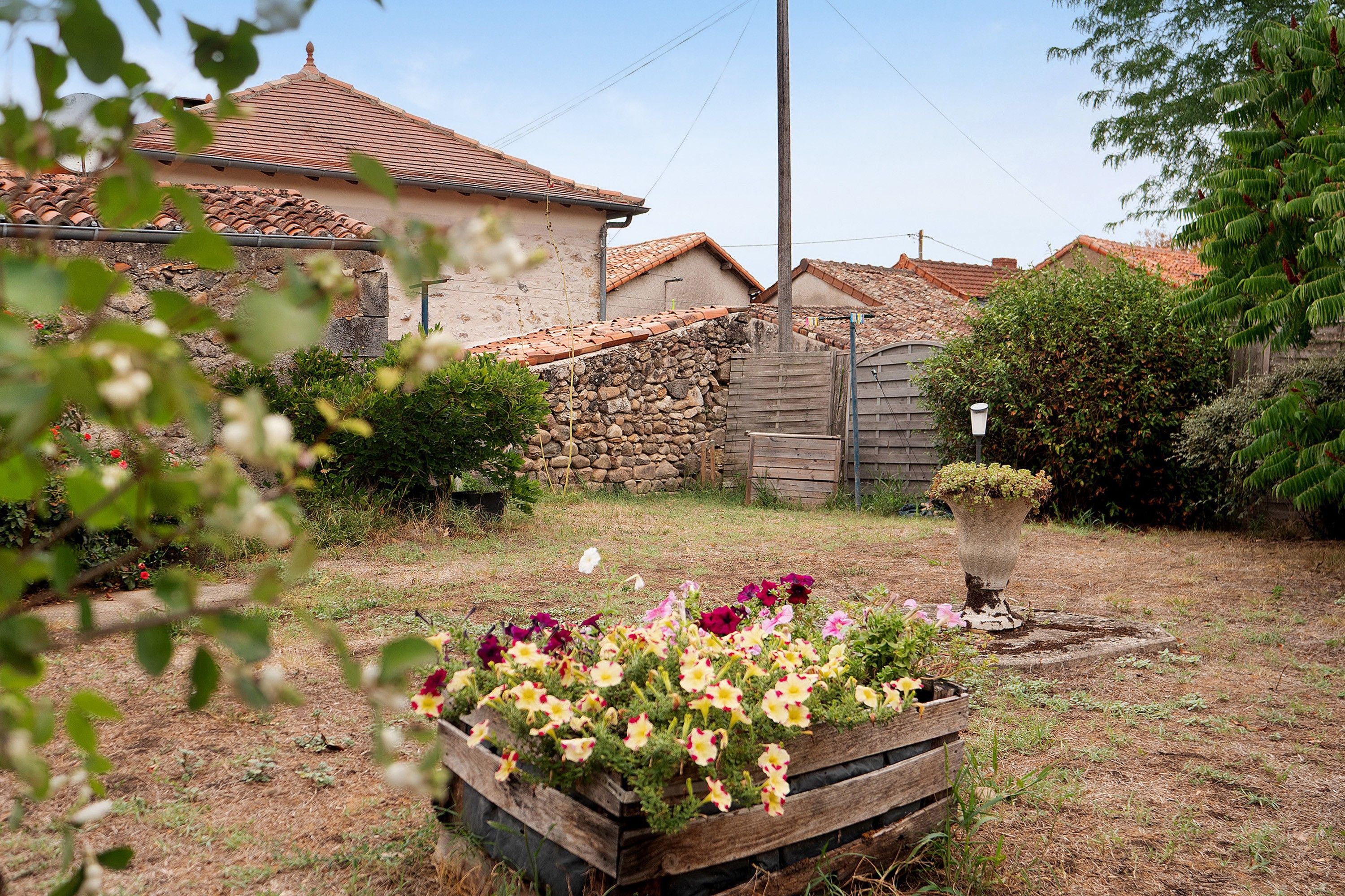 Un jardin avec un bac à fleurs en bois, un mur de pierre et une maison avec un toit en tuiles.