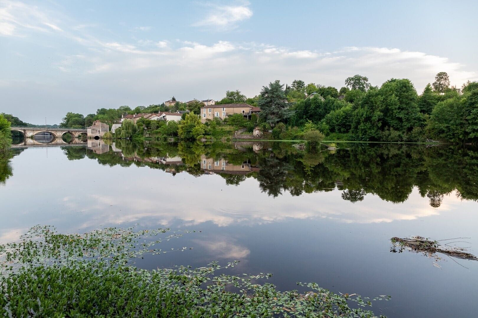 Ein ruhiger Fluss mit einer Brücke, Gebäuden und üppigem Grün.