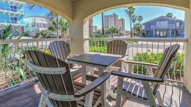 A covered outdoor seating area with Adirondack chairs and a table, overlooking a street with buildings and palm trees. - Athena Ares Seaside Condo (Floor 2) Pool • Hot Tub (South Padre Island)