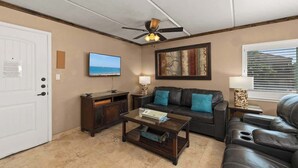 A living room with a ceiling fan, a television, a sofa, a coffee table, and a window with blinds. - Athena Ares Seaside Condo (Floor 2) Pool • Hot Tub (South Padre Island)