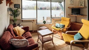 Un salon comprenant un canapé, des fauteuils, une petite table, une fenêtre donnant sur un plan d’eau et un radiateur.