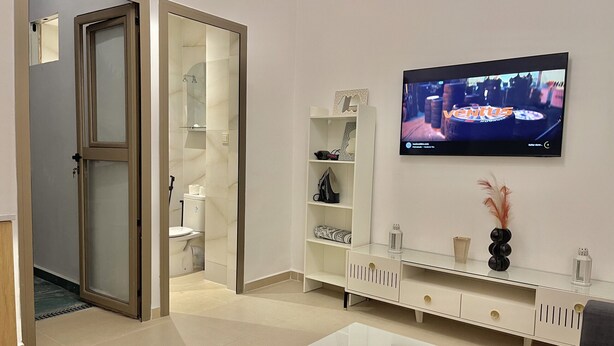 Una sala moderna con una mesa de centro de vidrio, un televisor de pantalla plana montado en la pared, una consola blanca con objetos decorativos y una puerta corrediza de vidrio que da al baño.