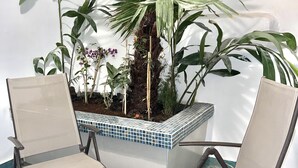 Un área de estar con dos sillas y una mesa pequeña, rodeada de plantas en macetas y una jardinera de azulejos.