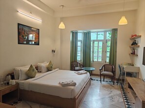 A hotel room with a bed, bedside table, chairs, a desk, a window with curtains, a painting on the wall, and a shelf with items.