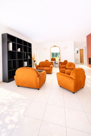 A modern living room with a black bookshelf, orange sofas, a round mirror, and a tiled floor. - VILLA DE STANDING DOMAINE DE MASSANE - Golf (Baillargues)
