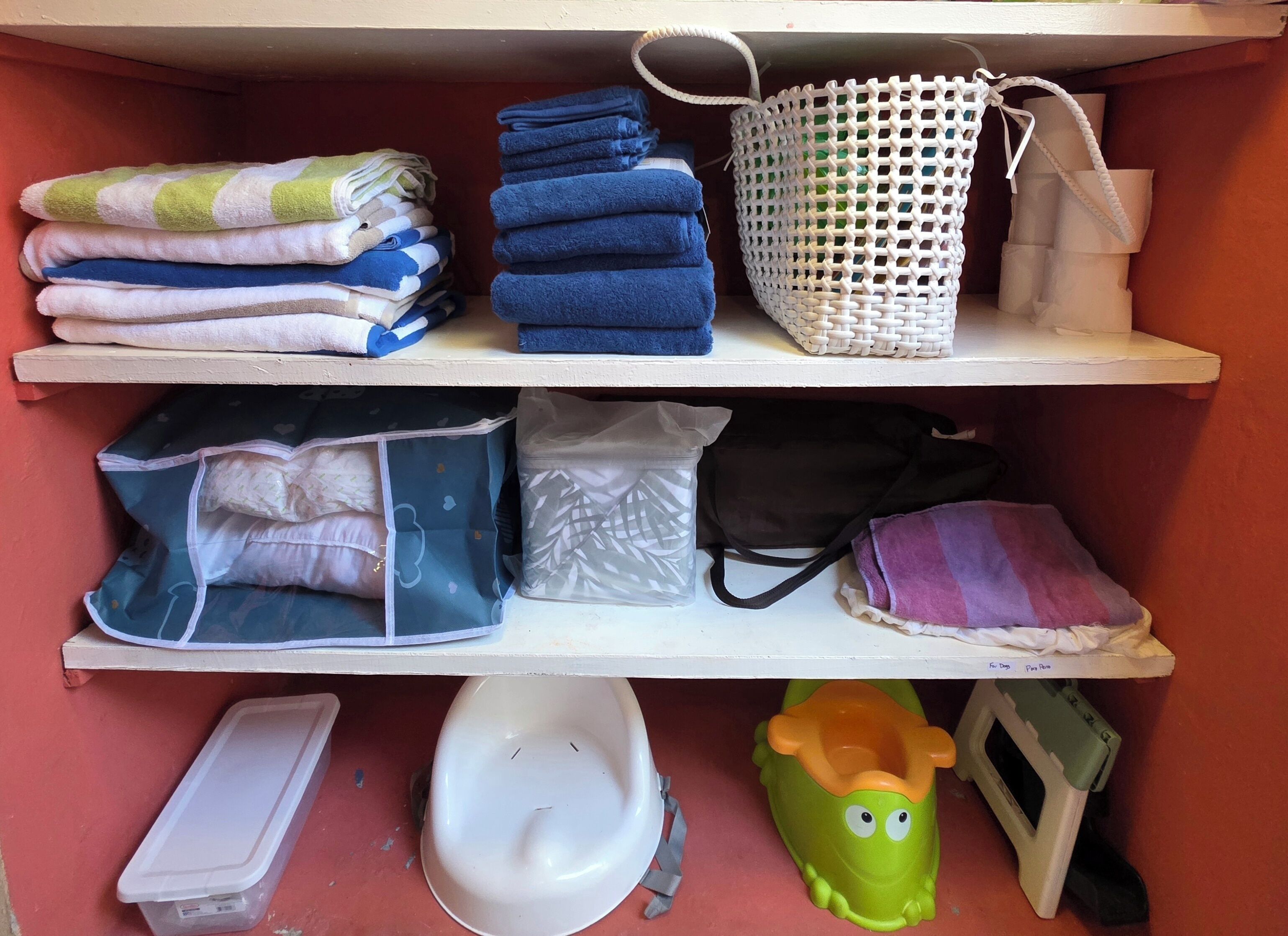 A shelf with folded towels, a woven basket, plastic bags, a black bag, and a toy.