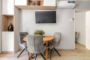 Ein moderner Essbereich mit einem runden Holztisch, vier grau gepolsterten Stühlen, einer Topfpflanze, einem flachbildfernseher an der Wand und einer Klimaanlage.