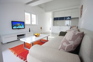 Ein modernes Wohnzimmer mit Flachbildfernseher, einem Esstisch, einer Couch und einer Küche im Hintergrund.