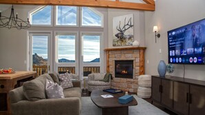 Una sala de estar acogedora con chimenea de piedra, ventanas amplias, un televisor y vistas a las montañas.