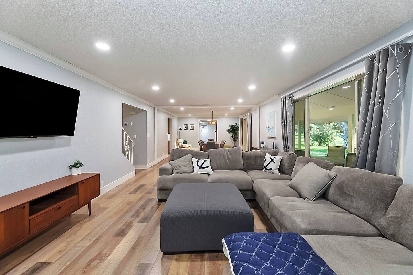 A modern living room with a sectional sofa, a flat-screen TV, wooden flooring, and large windows.