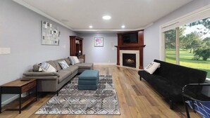 A living room with a sectional sofa, a black armchair, a fireplace, and a large window offering a view of a green landscape. - 5BR Boise Retreat | Perfect for Groups or Families (Boise)