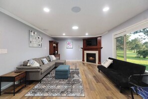 A living room with a sectional sofa, a black armchair, a fireplace, and a large window offering a view of a green landscape. - 5BR Boise Retreat | Perfect for Groups or Families (Boise)