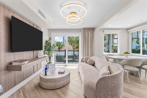 Ein modernes Wohnzimmer mit Flachbildfernseher, rundem Couchtisch, beiger Couch und Essbereich mit weißem Tisch und Stühlen.