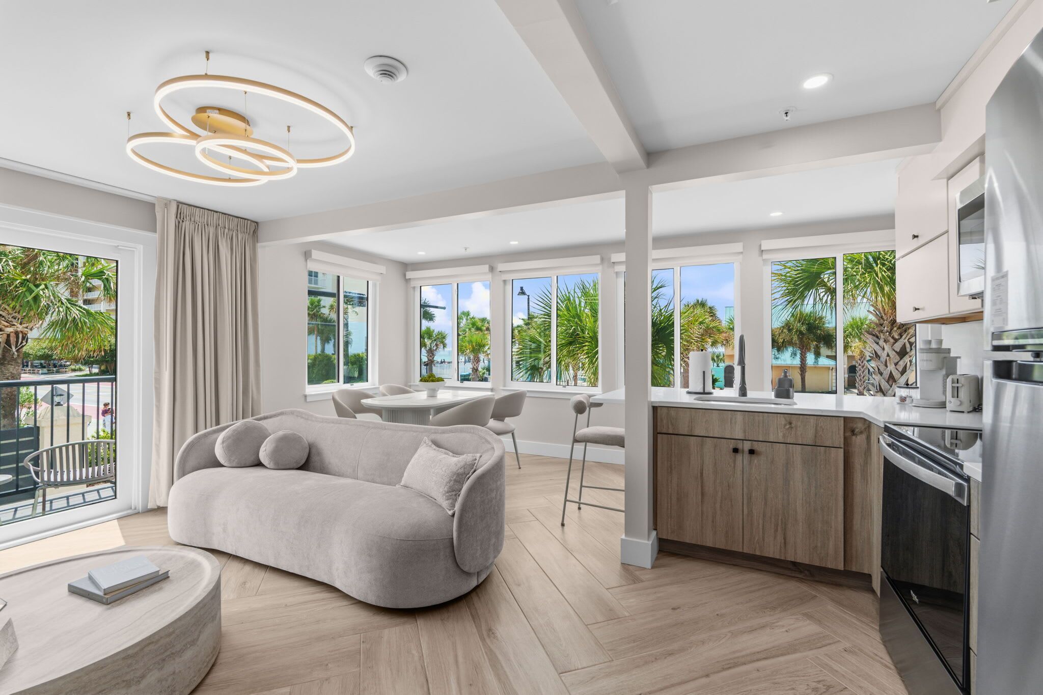 A modern living room with a sofa, a dining area, and a kitchen with a view of palm trees.