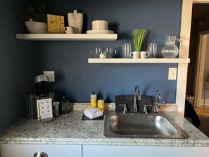 Una cocina con encimera de granito, pileta de acero inoxidable y muebles blancos. Hay dos repisas sobre la encimera con diferentes utensilios de cocina.