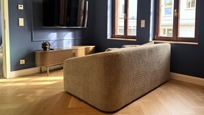 Una habitación con una televisión de pantalla grande montada en la pared, una mesita consola de madera, un sofá color beige y una ventana con vistas a un edificio.
