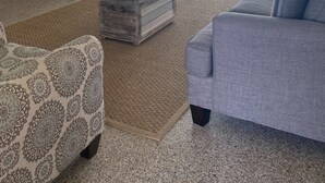 Una sala de estar con un sofá gris, un sillón estampado y un reposapiés. Se ve una cocina con gabinetes blancos y un televisor de pantalla plana al fondo.