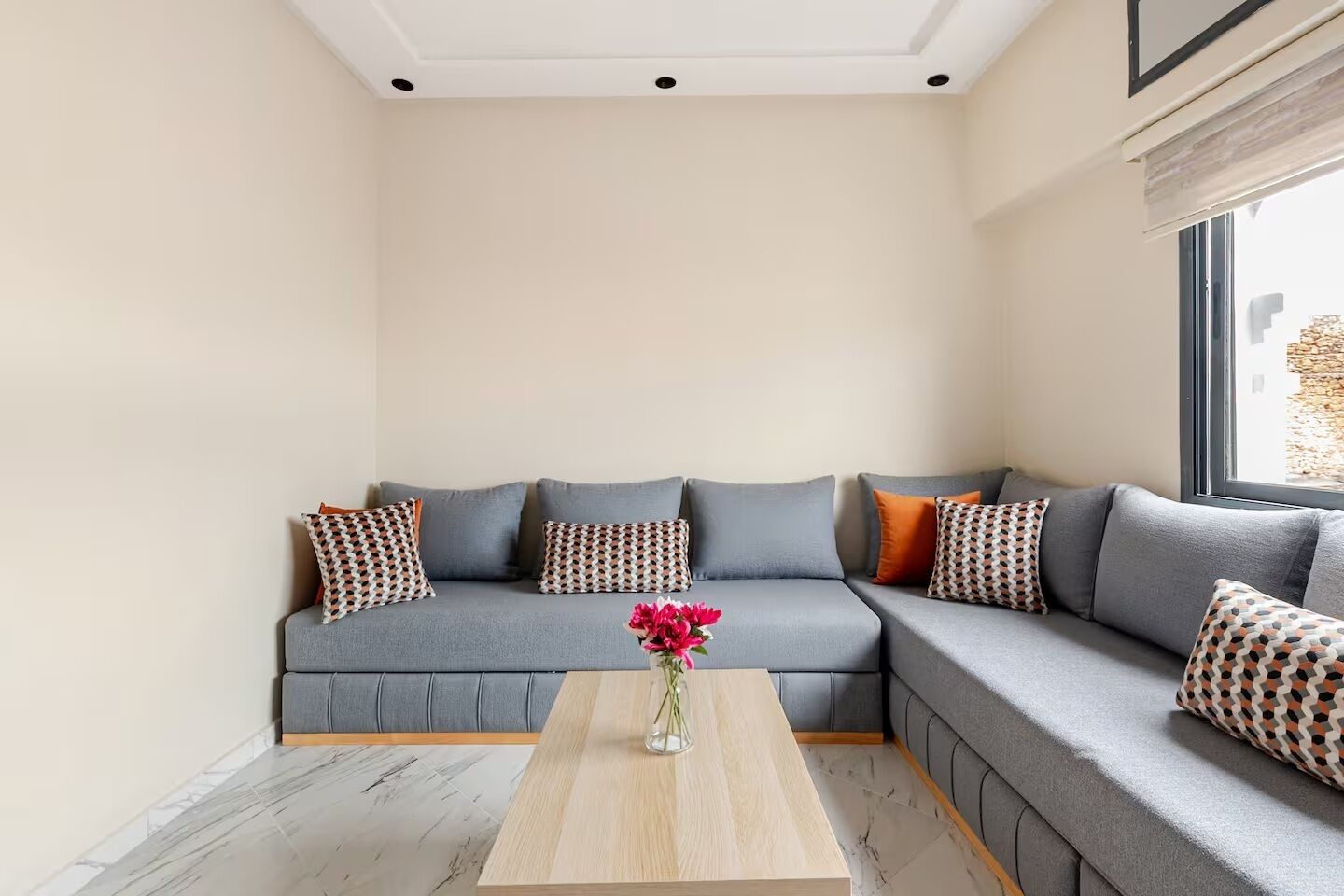 Un salon moderne avec un canapé d’angle gris, des coussins décoratifs à motifs, une table basse en bois et un vase rempli de fleurs.