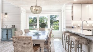 A modern kitchen with white cabinetry, a central island, and a dining area with wicker chairs and a table. - Lakefront Luxury • Golf Cart Included (Miramar Beach)