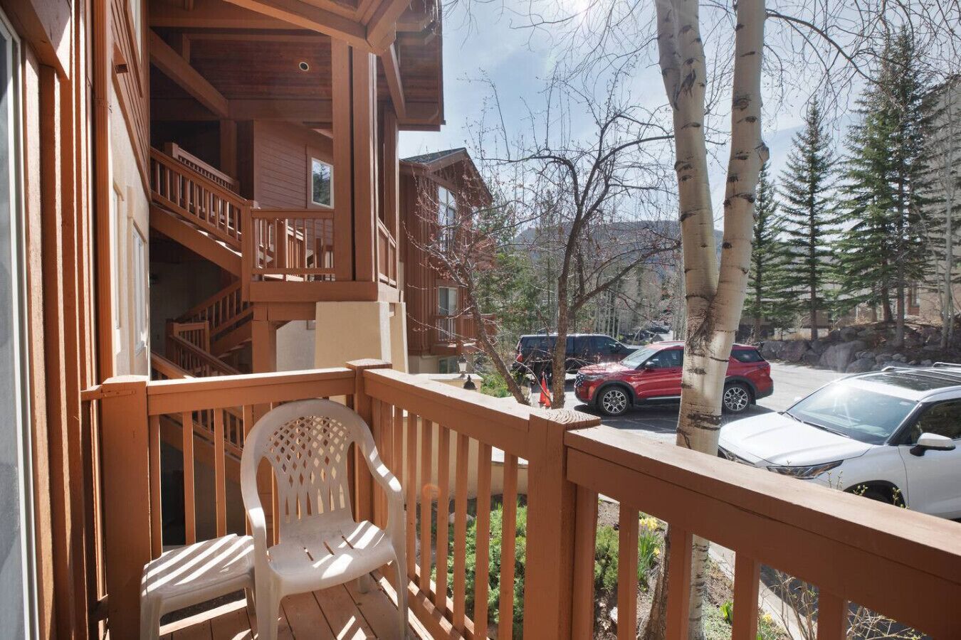 Un balcon en bois avec une chaise, donnant sur des voitures garées et une zone boisée.