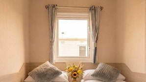 Una habitación con dos camas individuales, una ventana con cortinas, una mesita con un florero y una estantería con libros.