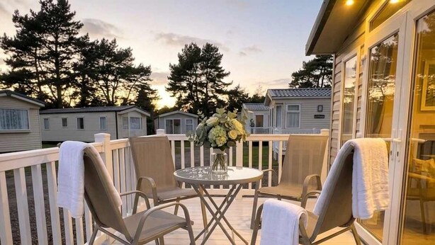 Een terras met een ronde tafel en vier stoelen, een vaas met bloemen, en een uitzicht op andere stacaravans op de achtergrond.