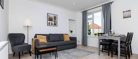 A modern living room with a sofa, a dining table with chairs, a lamp, and a window with curtains.