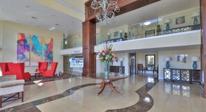 A lobby with a glass coffee table, red chairs, a chandelier, and a balcony with artwork. - Oceanfront & River View 3BD/2BA @ Grand Coquina 501 (Daytona Beach Shores)