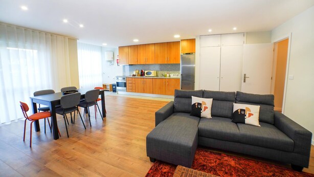 Uma sala de estar moderna com um sofá cinzento, uma mesa de jantar com cadeiras, uma área de cozinha com armários de madeira e um tapete vermelho.