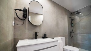 Casa de banho moderna com toucador branco, espelho redondo, secador de cabelo, sanita e chuveiro com parede de vidro.