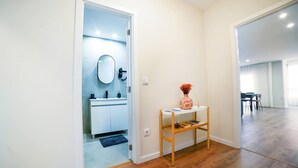Um corredor com chão de madeira, uma porta branca que dá para um banheiro e uma pequena mesa de madeira com um vaso.