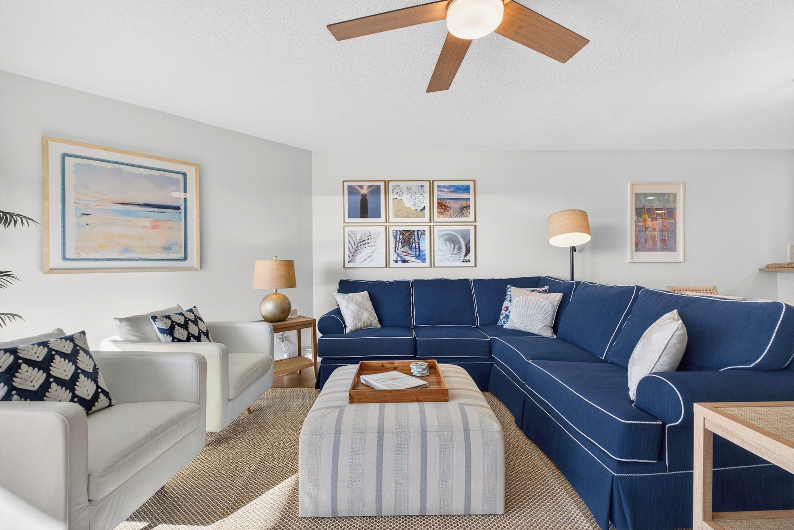A living room with a blue sectional sofa, a white armchair, a striped ottoman, a ceiling fan, and framed artwork on the walls.