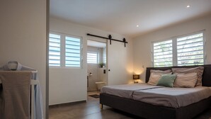 A bedroom with a bed, a nightstand, a lamp, a window, and a door leading to a bathroom. - Aqua Vista, Oceanview Villa with infinity pool Coral Estate close to the beach! (Sint Willibrordus)