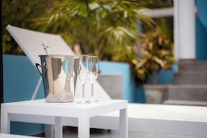Ein weißer Tisch mit einem silbernen Eimer und zwei Gläsern, vor einem blauen Stuhl und Grünpflanzen.
