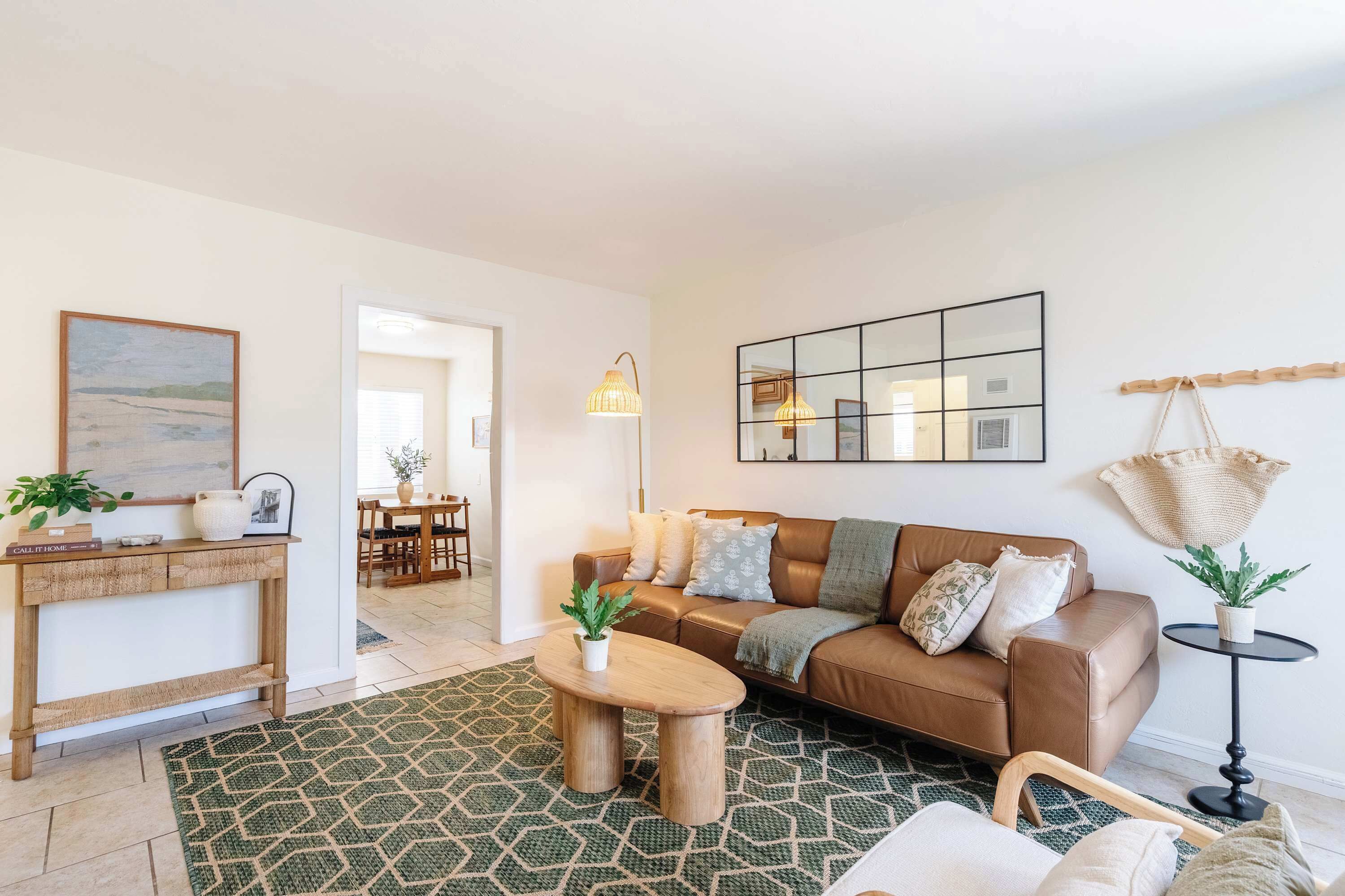 Een woonkamer met een bruine leren bank, een houten salontafel, een vloerkleed met dessin, een eethoek met tafel en stoelen, en een hangplant.