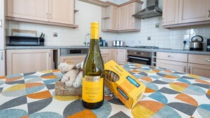 Uma garrafa de Sauvignon Blanc FirstCAPE da África do Sul, uma caixa de chocolate Moores e um ursinho de pelúcia sobre uma mesa em uma cozinha com armários de madeira clara e toalha de mesa estampada.