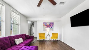 Uma sala de estar moderna com um sofá roxo, cadeiras amarelas, uma TV fixada na parede e um ventilador de teto.