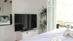 Ein Schlafzimmer mit Bett, Wandfernseher, Fenster und einer Zimmerpflanze.