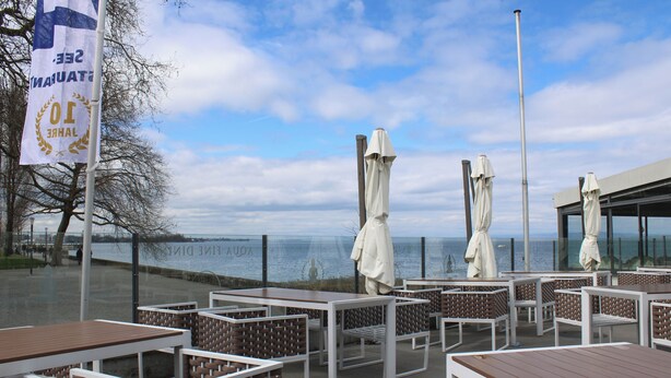 Terrasse mit Tischen und Stühlen, einer Flagge und Blick auf das Wasser.