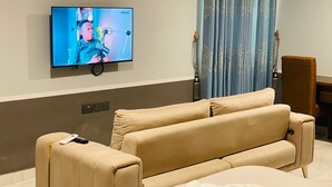 A hotel room with a beige sofa, a television mounted on the wall, a small table, and a chair. - SNL Mountain View Hotel (Akosombo)