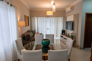 A modern living room with a glass dining table, white chairs, a sofa, a TV, and a lamp. - Casa c Churrasqueira a 350m da Praia de Massaguaçu (Caraguatatuba)