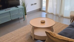 Un salon moderne avec un canapé, une table basse ronde, un téléviseur posé sur un meuble et une grande fenêtre avec des rideaux.