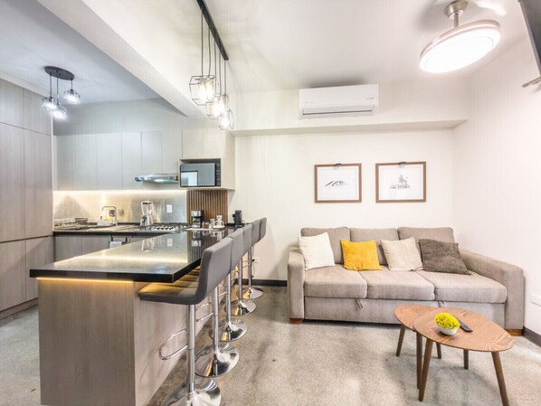 A modern kitchen with a central island, bar stools, a dining table, and a living area with a sofa and coffee table.