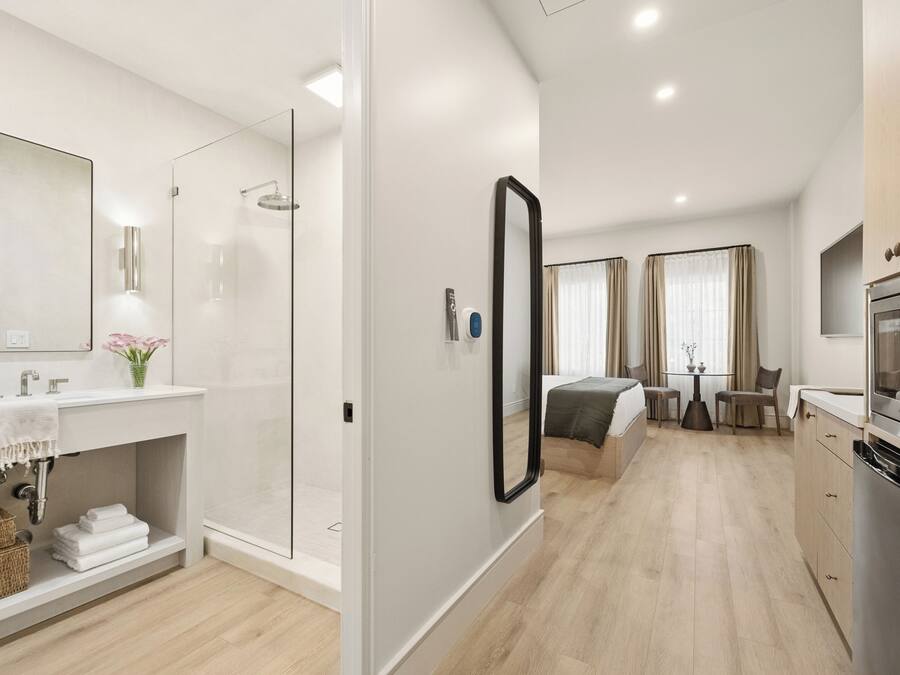 A modern hotel room with a shower, a large mirror, a bed, a dining area, and a kitchen.