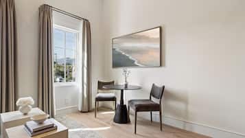 Ein modernes Wohnzimmer mit großem Fenster, einem runden Tisch, zwei Stühlen, einer Blumenvase und einem gerahmten Bild an der Wand.