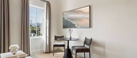 A modern living room with a large window, a round table, two chairs, a vase with flowers, and a framed picture on the wall.
