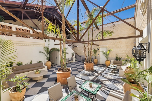 Une cour avec un sol à carreaux, des espaces aménagés pour s’asseoir, des plantes en pot et une structure de toit en métal.