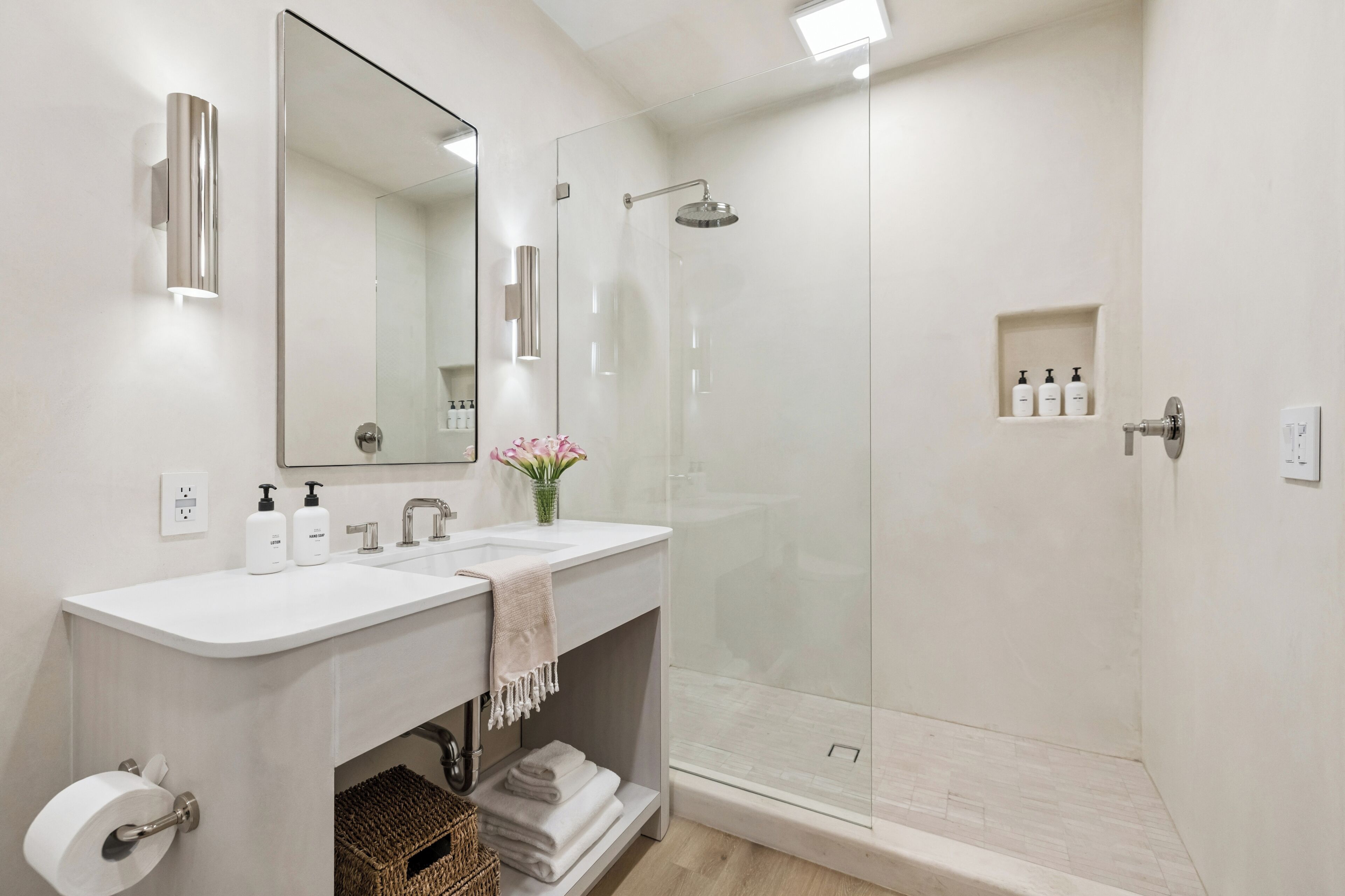 A modern bathroom with two sinks, a large mirror, a glass shower enclosure, and a small shelf with toiletries.