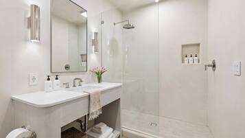A modern bathroom with two sinks, a large mirror, a glass shower enclosure, and a small shelf with toiletries.